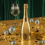 A shiny gold bottle of Lamborghini Extra Dry wine with a glass on a gold table. Scattered gold confetti, ribbons, and small silver disco balls around, against a dark green curtain background.