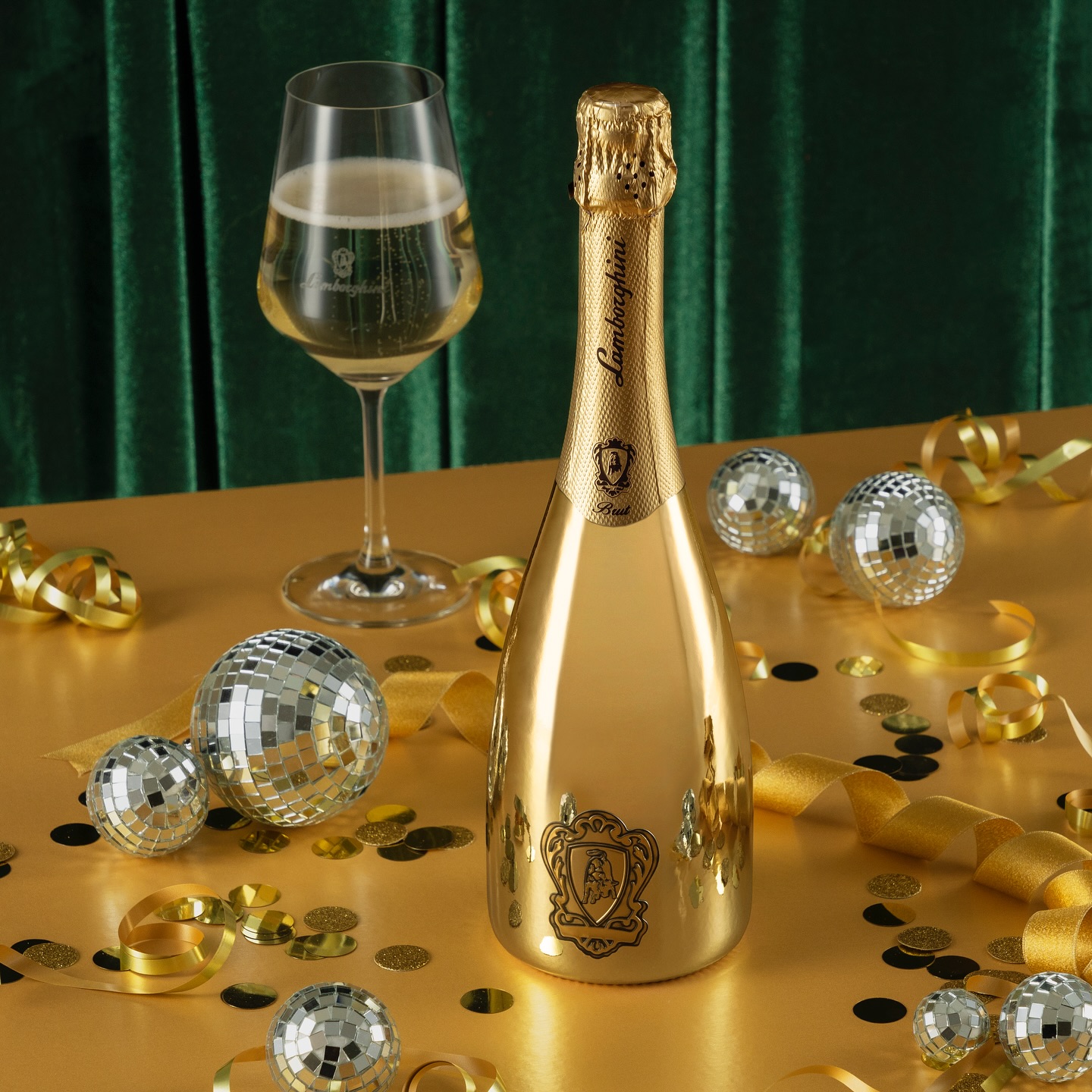 A shiny gold bottle of Lamborghini Extra Dry wine with a glass on a gold table. Scattered gold confetti, ribbons, and small silver disco balls around, against a dark green curtain background.