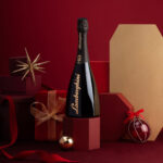 A bottle of Lamborghini Ottagonale Brut Gold wine displayed on burgundy pedestals among Christmas gifts tied with a gold ribbon, a red bauble, and gold stars on a deep red background.