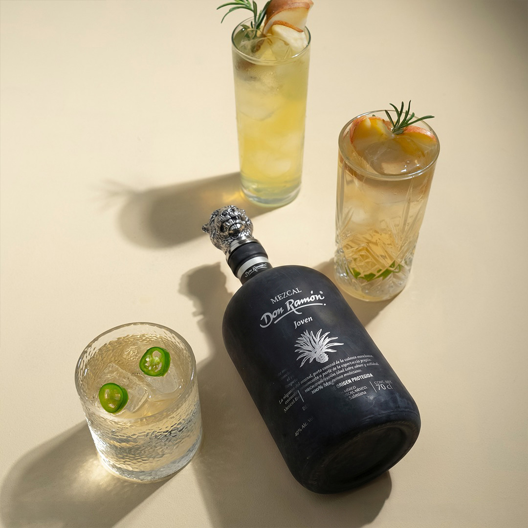 A black bottle of Mezcal Don Ramón Salmiana Joven lying on a light background. Around it are three glasses with different light cocktails, garnished with jalapeño slices, rosemary, and fruit.
