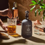 A bottle of Mezcal Don Ramón standing on a counter. Next to it, bartender's hands are visible stirring a brown cocktail with a long spoon in a glass mixing pitcher. Copper and steel bar accessories in the background.