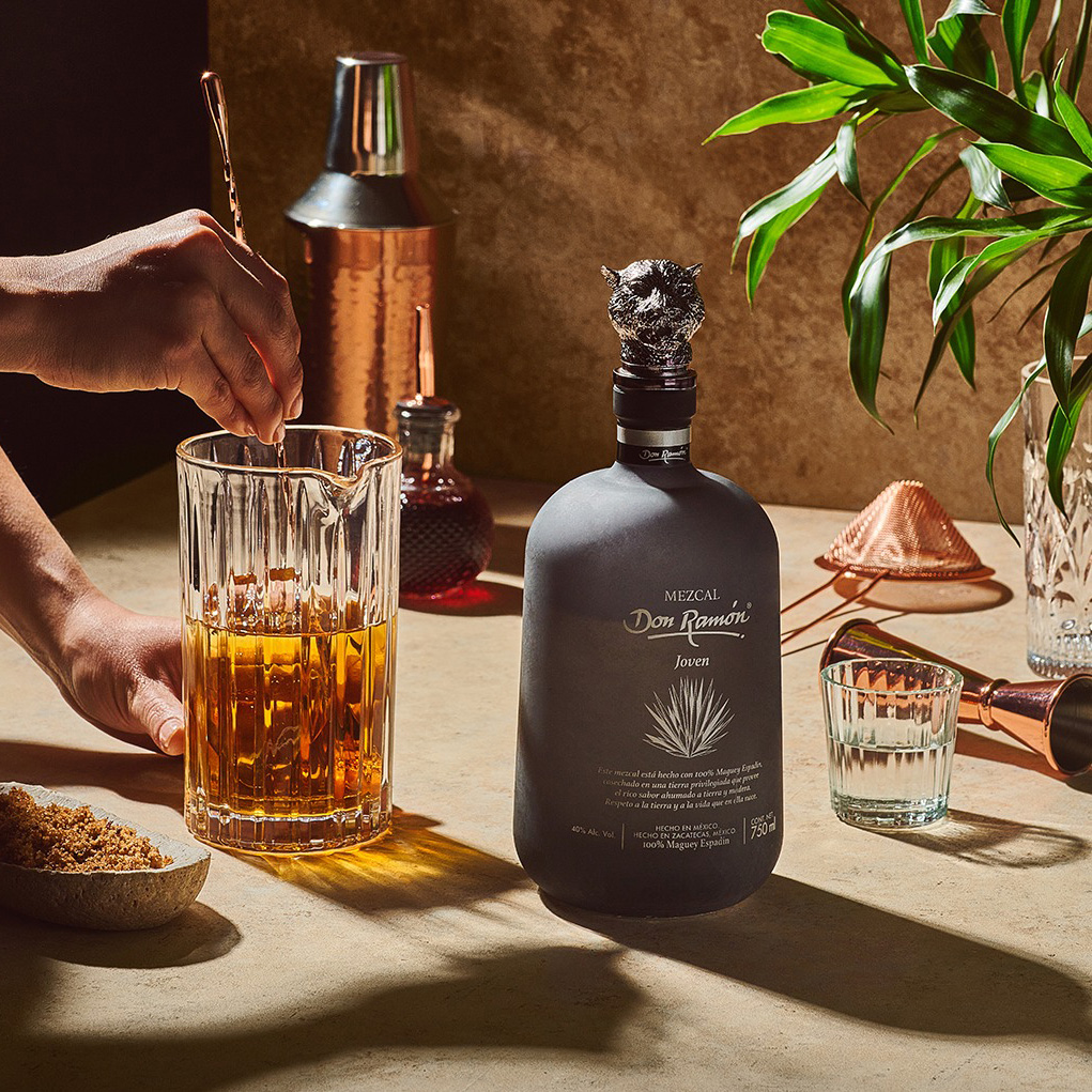 A bottle of Mezcal Don Ramón standing on a counter. Next to it, bartender's hands are visible stirring a brown cocktail with a long spoon in a glass mixing pitcher. Copper and steel bar accessories in the background.