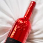 Close-up of a glossy red metallic bottle of Lamborghini Luxe Rosso wine with an embossed shield logo, lying on crumpled white sheets