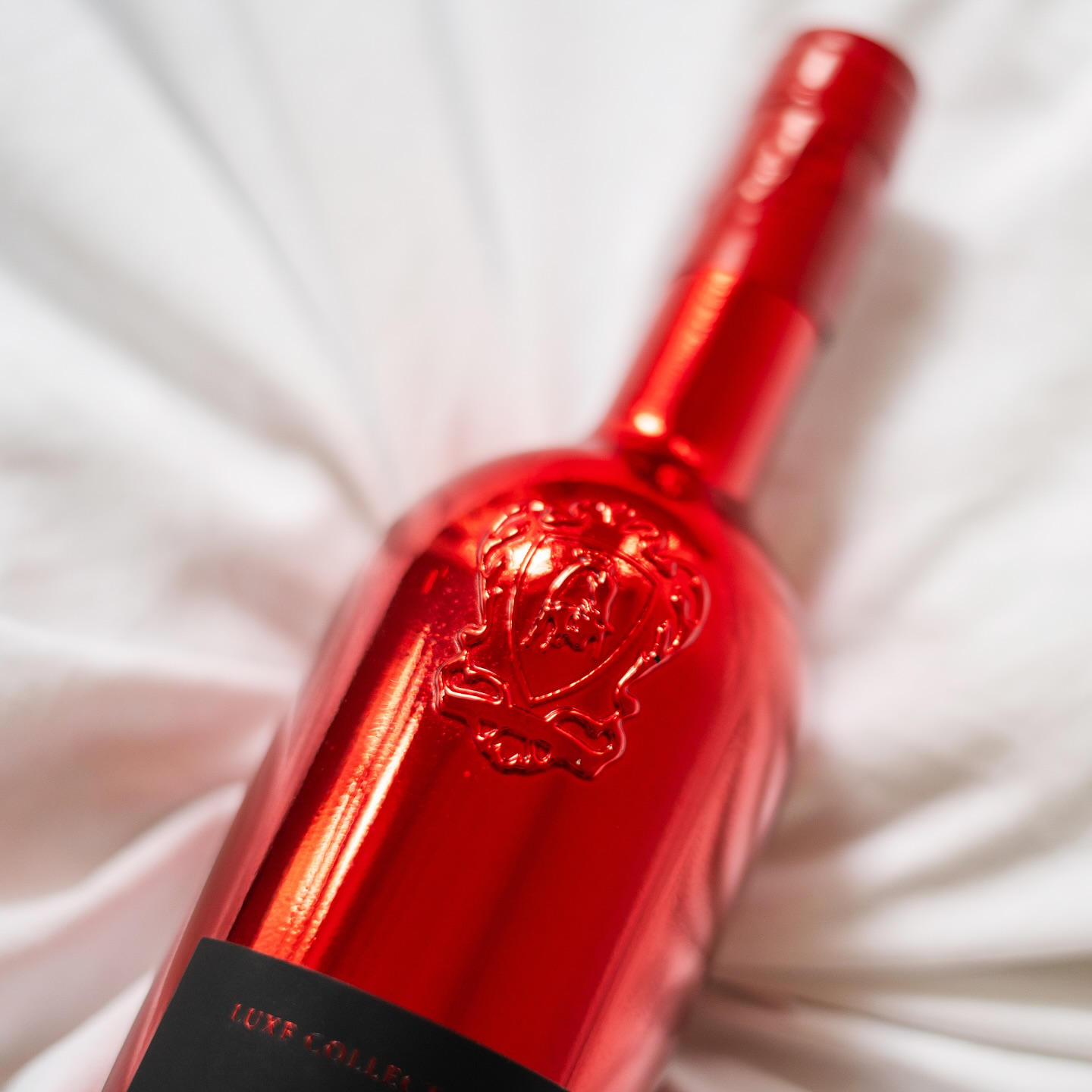 Close-up of a glossy red metallic bottle of Lamborghini Luxe Rosso wine with an embossed shield logo, lying on crumpled white sheets