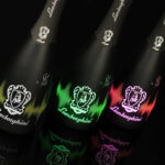 black bottles of Lamborghini DJ Luminous Brut wine lined up in the dark. Each bottle has an illuminated logo in a different color: yellow, green, pink/red. Reflections are visible on a glossy surface.
