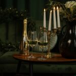 A gold bottle of Lamborghini Extra Dry Gold wine on a wooden table with two glasses, next to lit candles in a brass candelabra against a dark green background.