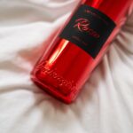 Macro shot of the red metallic Lamborghini Luxe Rosso wine bottle on white bedding, showing the black label with red lettering and the elegant embossed Lamborghini name at the bottom