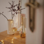 Cozy, home arrangement: a gold bottle of Lamborghini Extra Dry wine, a glass, and a vase with winter branches on a wooden dresser, decorated with warm fairy lights (LEDs).