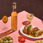 A bottle of Tequila Don Ramon Anejo on a pink tabletop surrounded by cocktails, two meat sandwiches, and a bowl of snacks.