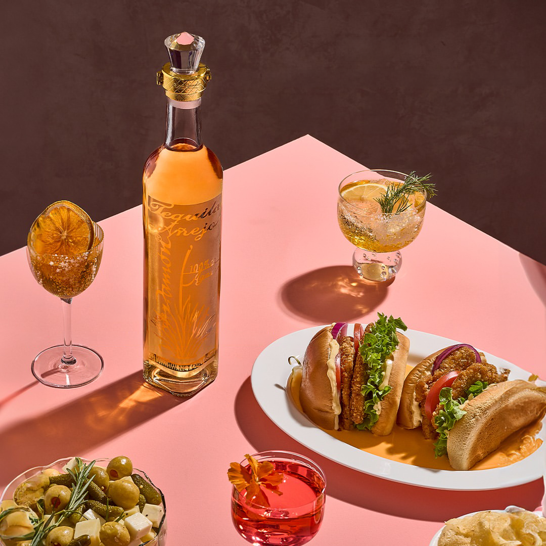 A bottle of Tequila Don Ramon Anejo on a pink tabletop surrounded by cocktails, two meat sandwiches, and a bowl of snacks.