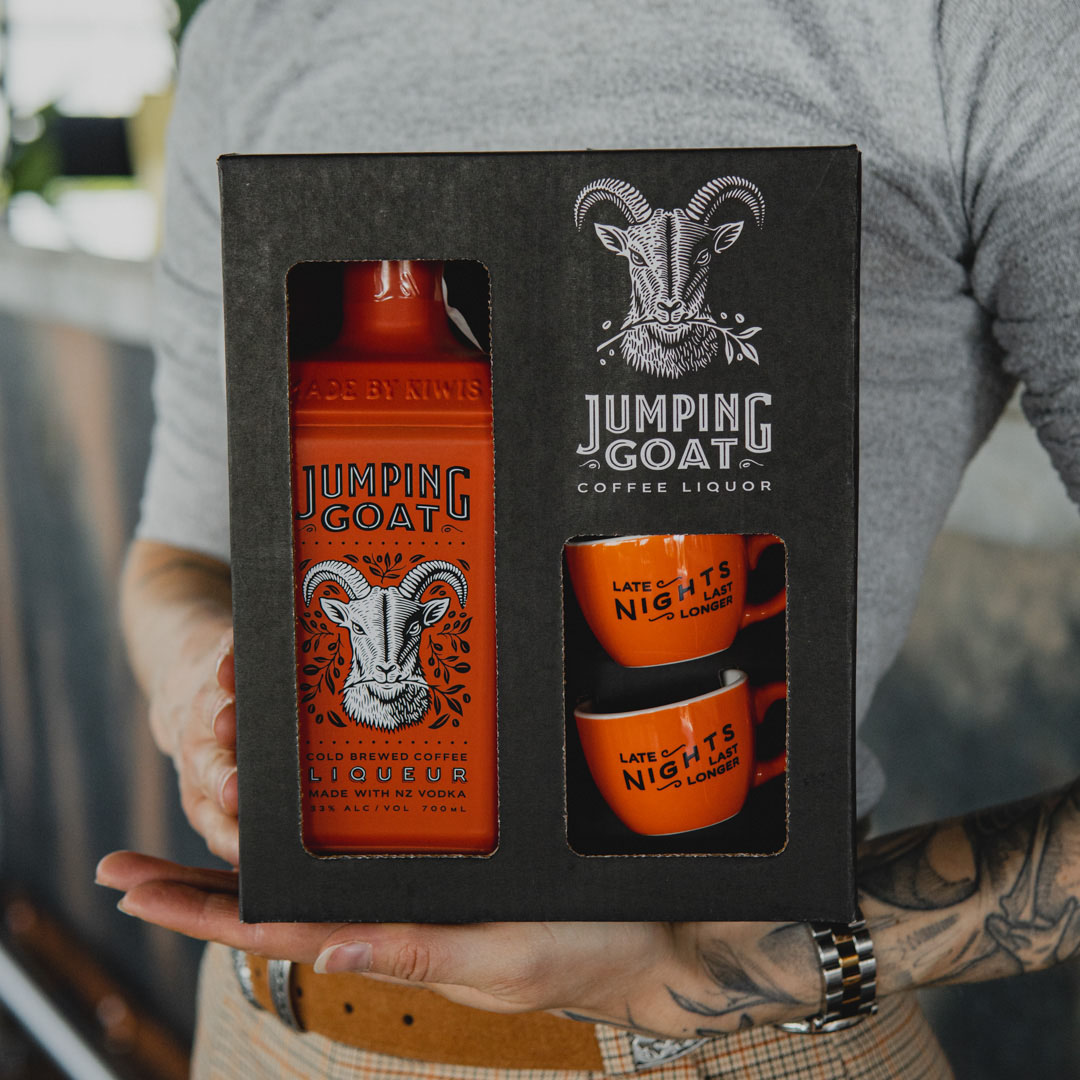 Osoba w szarym swetrze trzymająca z przodu czarne pudełko zestawu prezentowego Jumping Goat z pomarańczową butelką likieru kawowego na bazie wódki i dwiema filiżankami