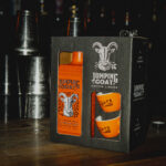 Czarny kartonowy zestaw prezentowy z pomarańczową butelką likieru kawowego Jumping Goat Vodka i dwiema filiżankami, stojący na drewnianym blacie baru. W ciemnym tle metalowe shakery barmańskie